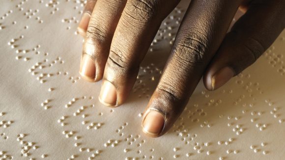 A close up photograph of someone's hand running over a sheet of braille.
