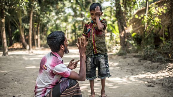 Polok stands and holds one hand over one eye. He looks down at his father who is sitting on the ground, holding up a high five to test Polok's vision.