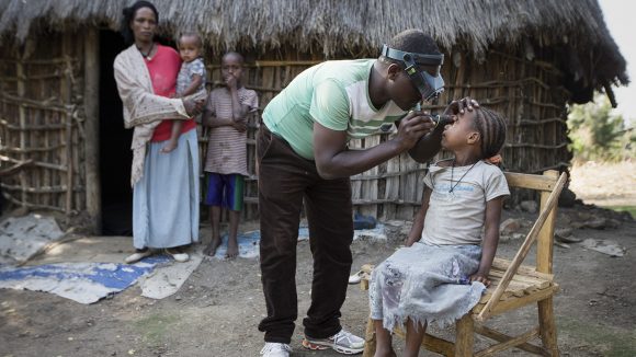A child is sitting on a wooden chair. A health worker is examining their eyes. Hibret and two other children are standing, watching in the background.