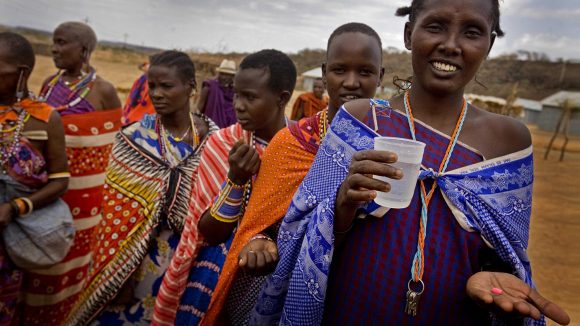 Six women wearing bright clothing stand outside. The women in the forefront is smiling, holding a pink pill in her hand and a glass of water in the other hand.