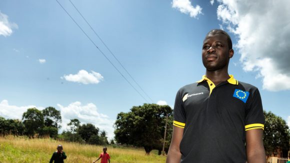 Atugonza stands proudly outside on a sunny day, wearing his Sightsavers-branded t-shirt.