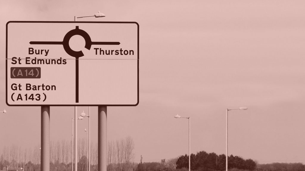 A road sign featuring the Transport font, with the words 'Bury St Edmunds (A14), Gt Barton (A143) and Thurston' plus a roundabout symbol.