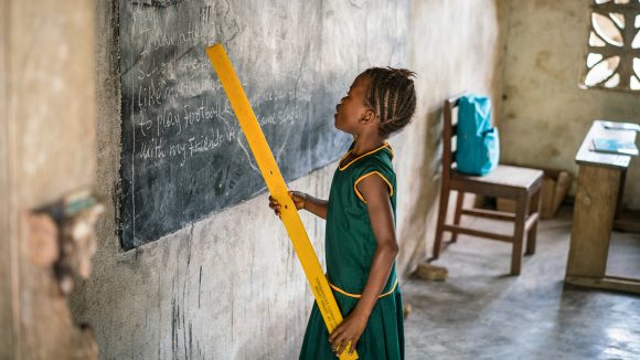 Aminata stands at the blackboard, holding a ruler to point out words.