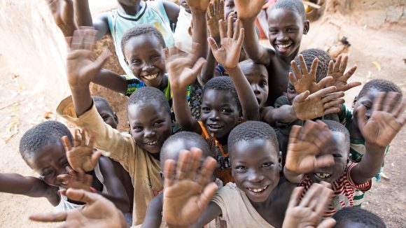 A group of children in Ghana smile and wave at the camera.