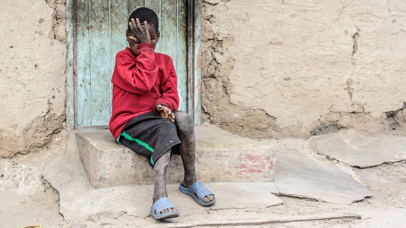 Mwiza sits outside his home, covering his eyes with his hand.