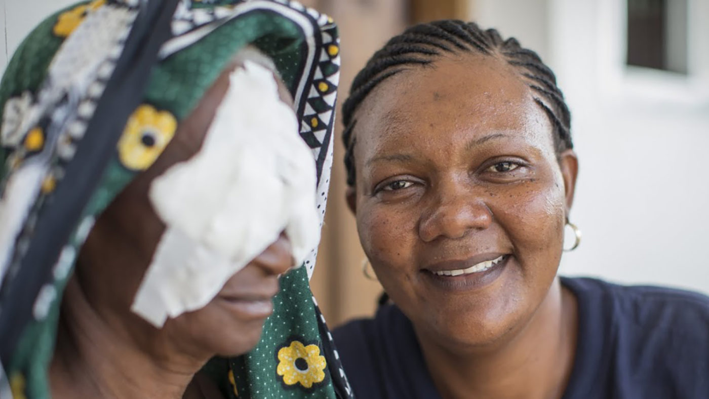 An eye care worker smiles at the camera next to a woman with bandages on her eyes.
