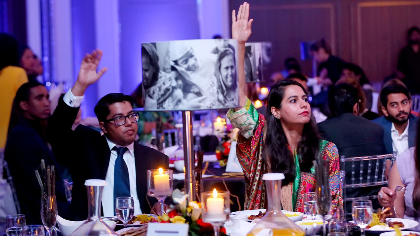 A couple sitting at an extravagant dinner table raise their hands in the air to bid during the auction.