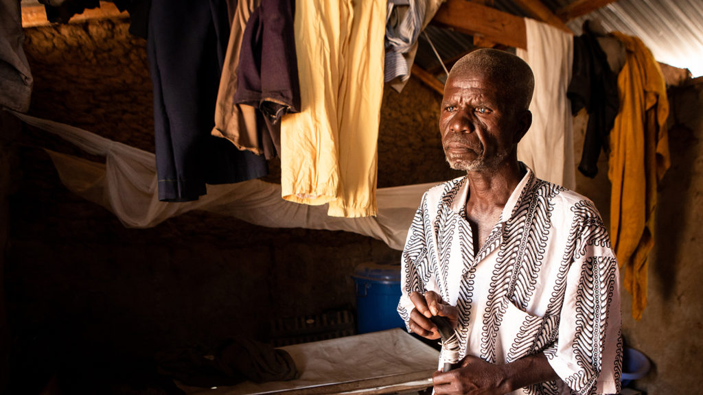 Isaac sits in his house, with clothing on a line behind him.