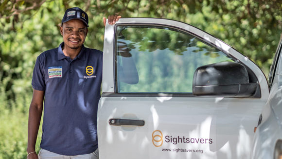An eye health worker wearing a purple t-shirt with the People's Postcode Lottery and Sightsavers logos, standing next to a van with the Sightsavers logo on the door.