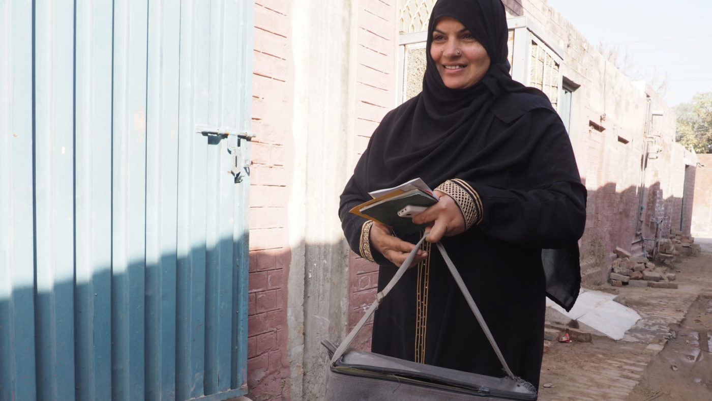 A woman wearing a black hijab smiles and walks along a brick-lined street.