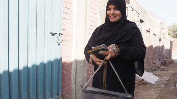 A woman wearing a black hijab smiles and walks along a brick-lined street.