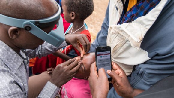 A Tropical Data trainee checks the eyes of a young child for signs of trachoma, while someone enters the data into the Tropical Data system on a mobile phone.
