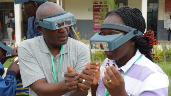 A male Tropical Data trainer shows female trainee how to identify what trachoma follicles would look like by using small, circular, red stickers with five white dots on their thumbs.