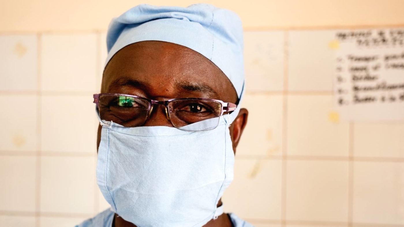 A trachoma surgeon stands for a portrait wearing his surgical scrubs and mask.