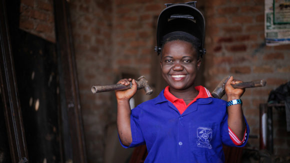 A woman wears a welding mask and carries tools.