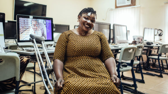A women with crutches sits next to computers in an office smiling.