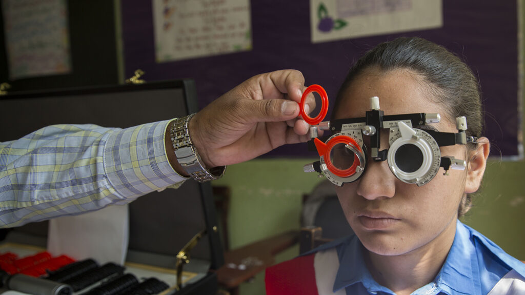 A school student has her eyes tested while wearing optical glasses: an eye health worker's hand is changing the lenses in the glasses.