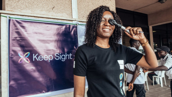 An eye health worker stood infront of a Keep Sight banner holds an eye examining instrument to her eye.