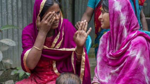 Shamima communicates with her sister. Her sister uses hand movements to get her point across. Both women are sitting down outside and wear bold, pink clothing.
