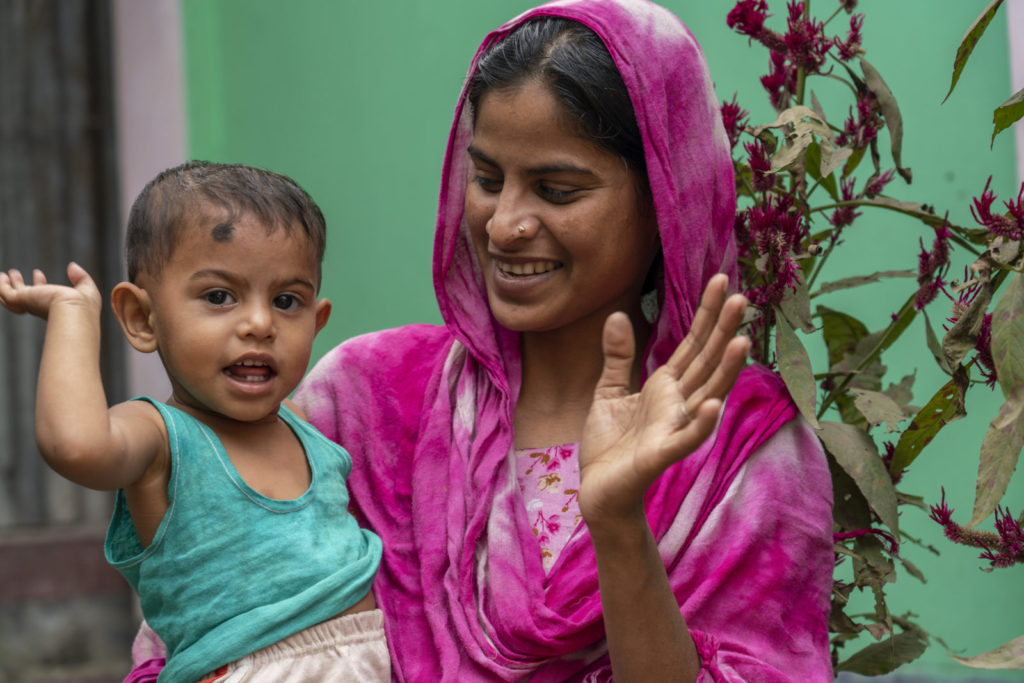 Shamima communicating with her young daughter via facial expressinons and hand signals. She wears a bright pink sari against a green backdrop.
