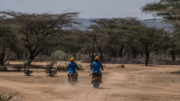 two volunteers ride motorbikes on a dirt road.