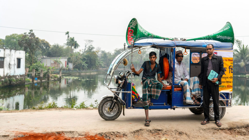 A colourful tuk-tuk with speakers on the roof and three men stood by it.