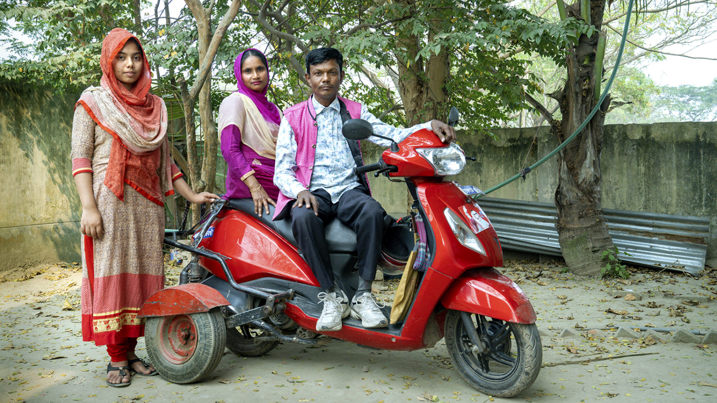 A man and wife sit on a motorbike with a young woman standing beside them.
