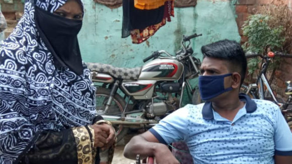 A woman stands next to a man sitting in a chair. Both wear face masks.