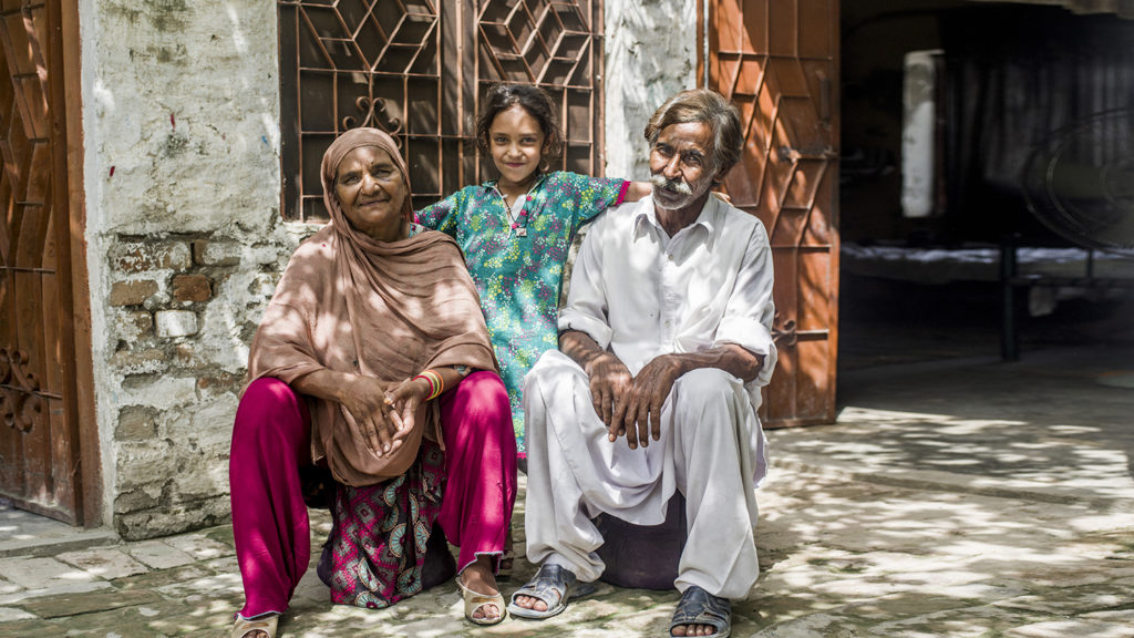 A mother and father sit on the ground with their daughter standing behind them.
