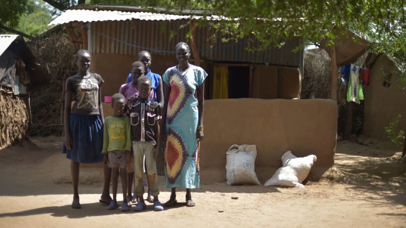Rebecca stands with her family outside her home, or refugee accommodation in Kenya.