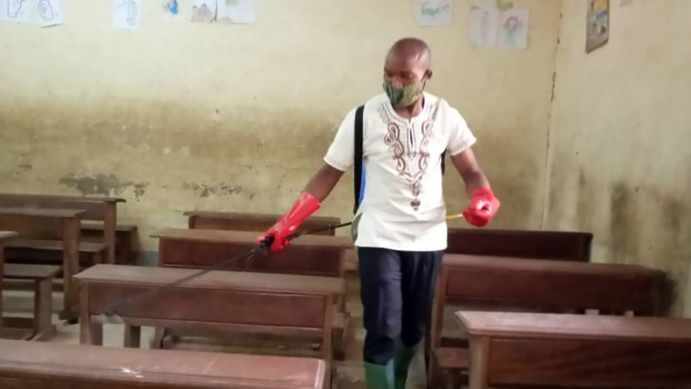 A man wearing a face mask and gloves sprays disinfectant in a classroom.