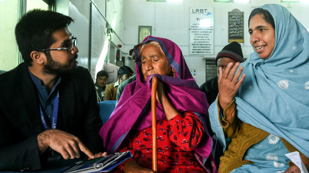 An elderly woman and a younger woman sit next to a man who holds a clipboard, in a hospital waiting area.