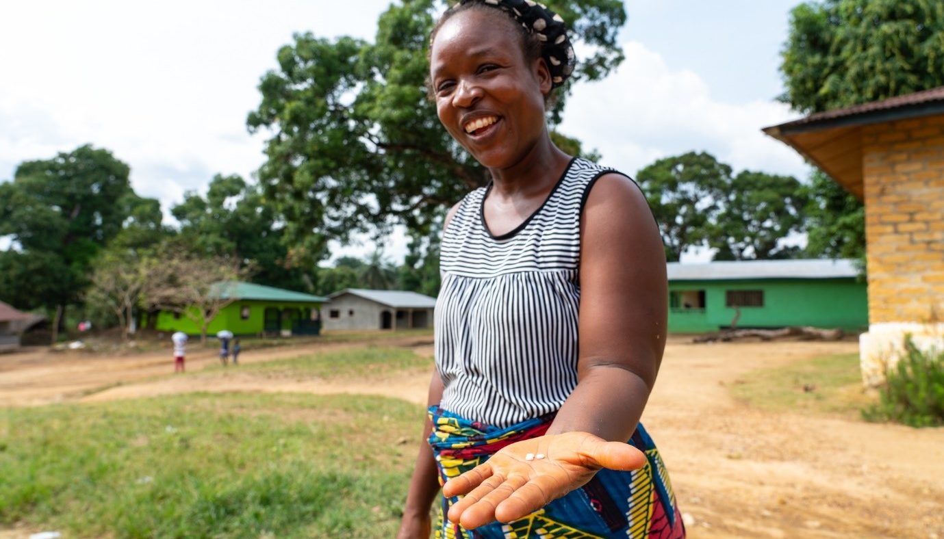 A woman stands outside smiling and showing the medication in her hand.