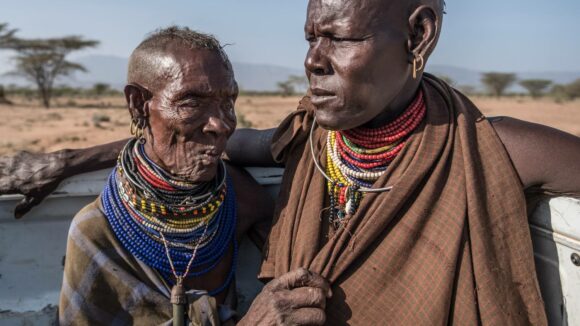 Two trachoma patients wearing traditional clothing standing next to a vehicle in a landscape in Kenya.