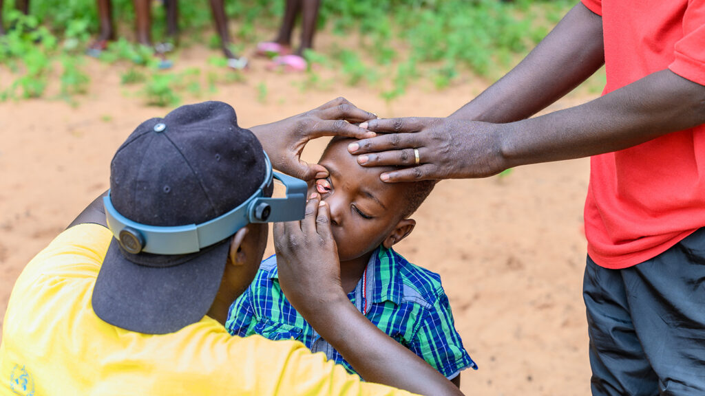 A medical nurse examines a child's eye while another adult puts a supportive hand on the child's head.