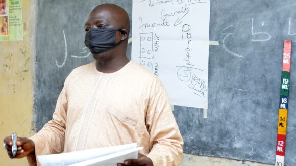 A man wearing a protective mask presents in front of a blackboard next to a dose poleivers training to antibiotics distributers in a classroom at Government Day Secondary School, Jigawa State