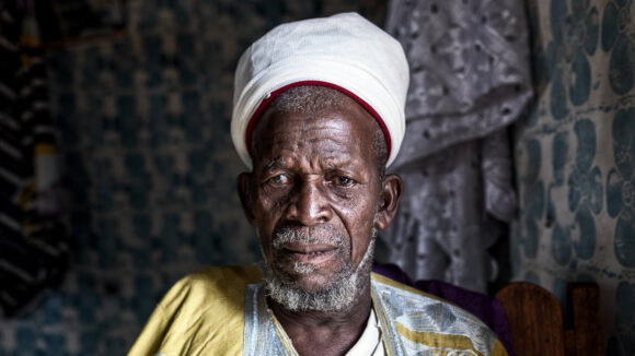 A man wearing traditional robes stands outside. He is looking down.