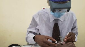 A man wearing a face mask, shield and optical loupes operates on the eye of a dummy