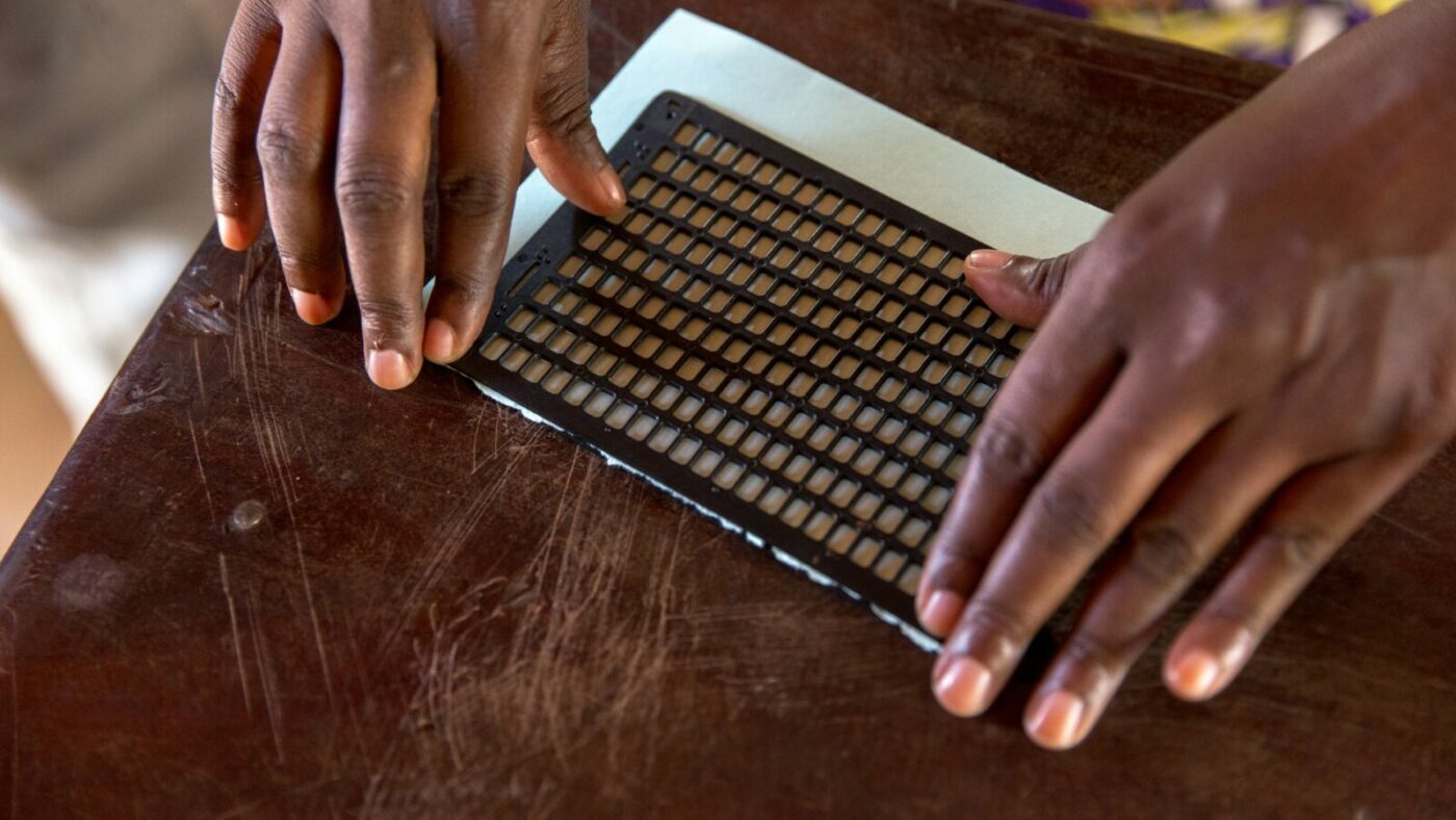 A student uses a braille machine, resting on a school desk. Only the student's hands are shown.