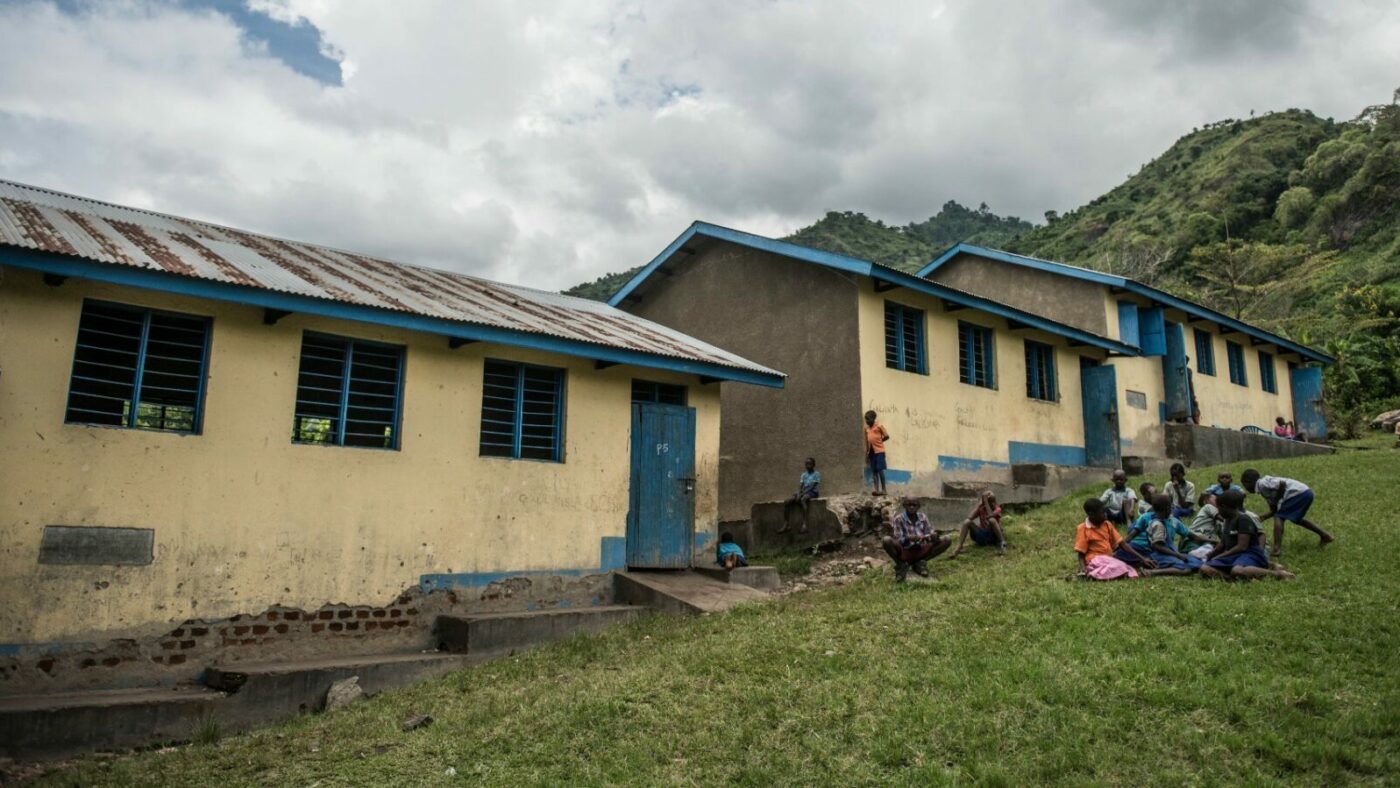 Children play in front of a long, low school building, with lush green hills in the background.