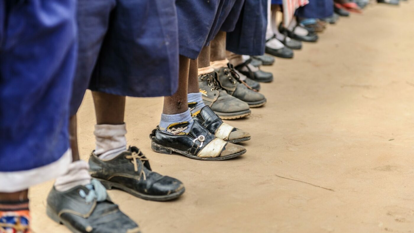 A line of schoolchildren stand in a school playing field. Only their feet and lower legs are shown. All are wearing blue trousers and black shoes.