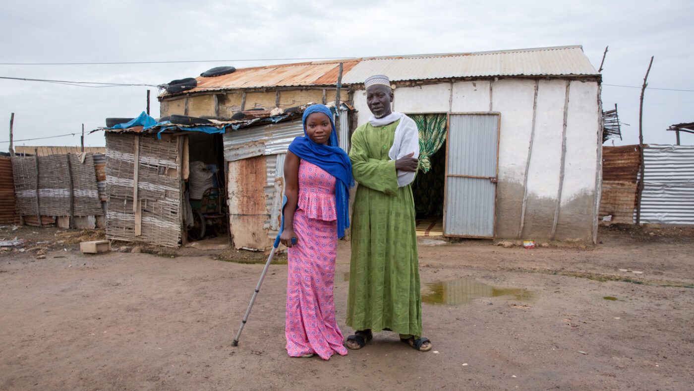 A man and woman stand together outside a dwelling. The woman is using a crutch.