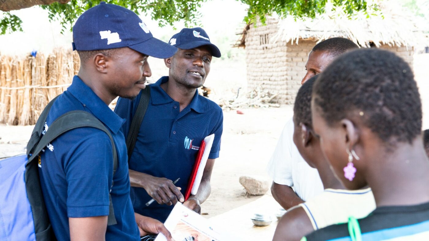 Two men wearing dark blue uniforms hand out paper surveys to a group of people.
