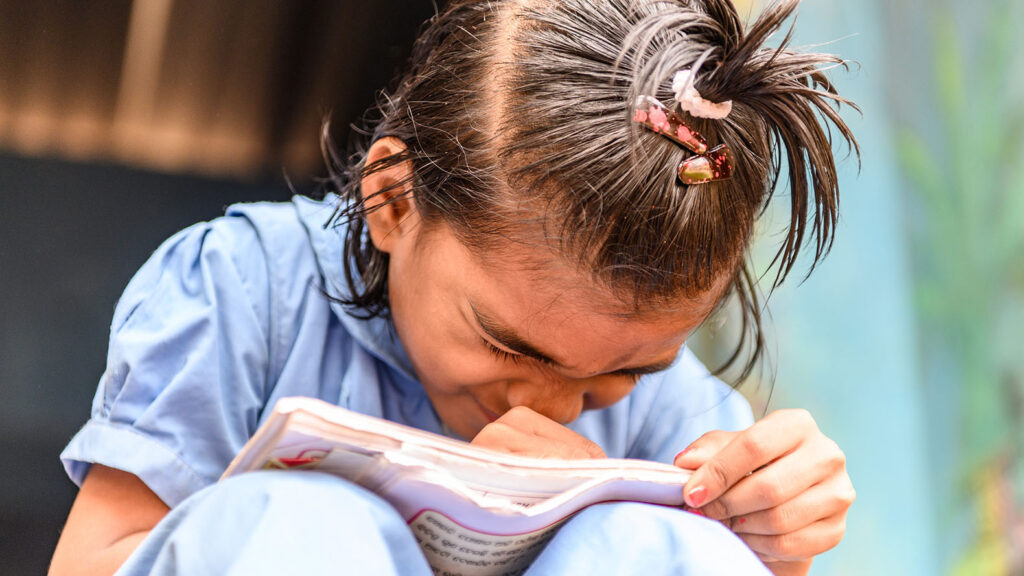 Archana is holding a book very close to her eyes and is squinting.