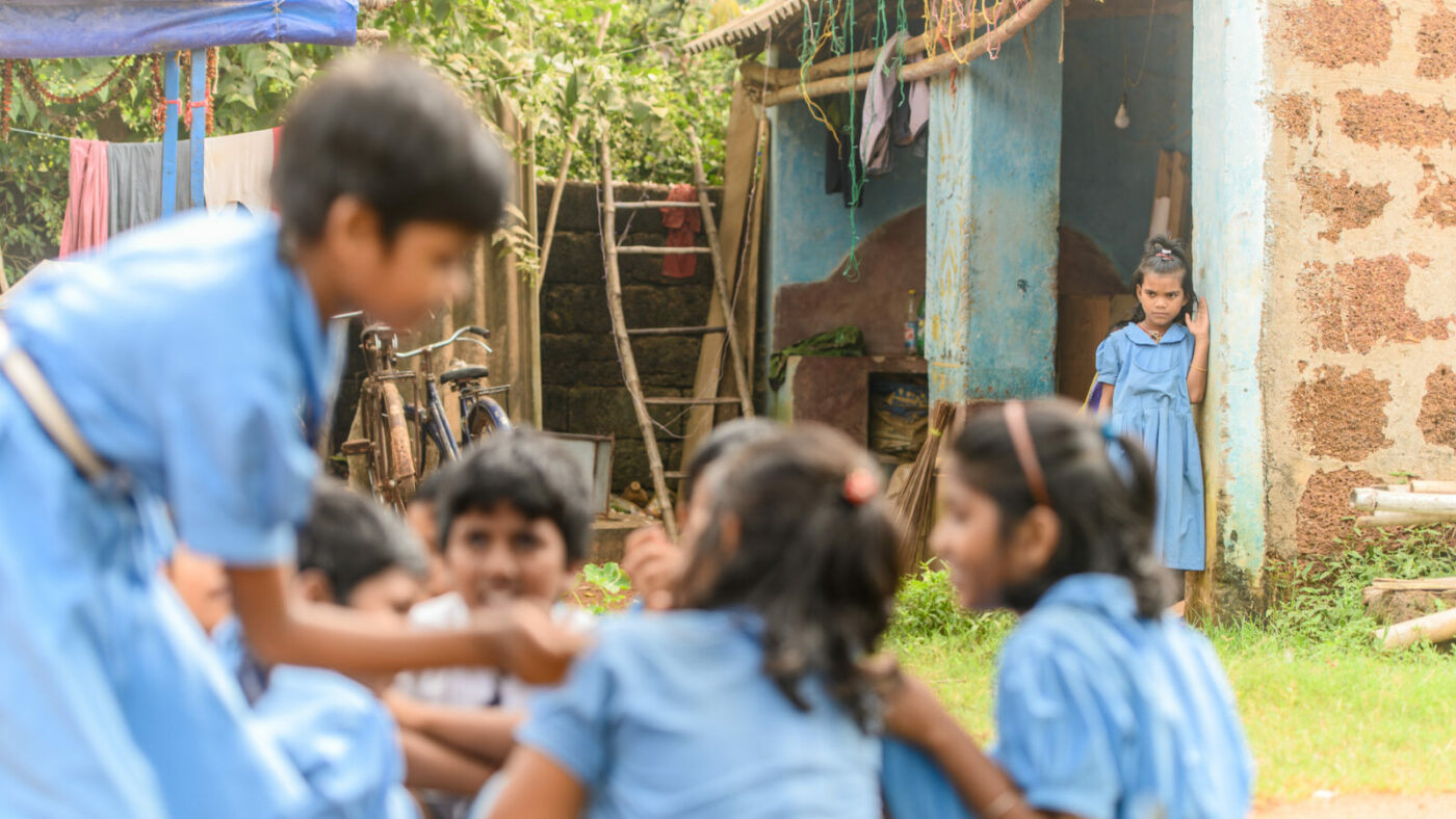 A young girl in school uniform looks at a group of friends playing, also in school uniform, from a wall.