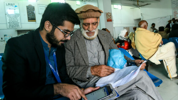 An older man talks to a younger man, who is holding a tablet computer. They are sitting in a hospital waiting room.