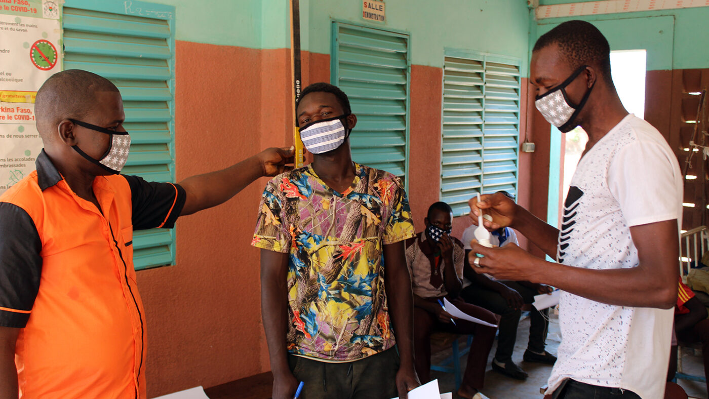 Two volunteers in masks are shown by a trainer how to measure a patient's height.