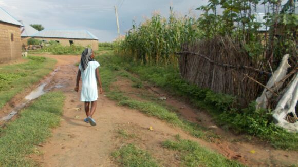 A girl, wearing a blue dress and a headscarf, walks away from the camera down a dusty track. The path she is walking on is next to a cornfield.