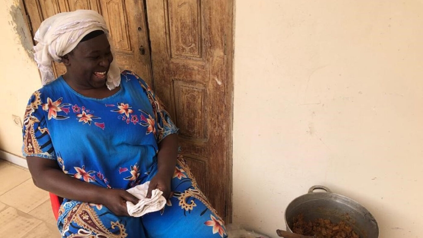 A lady smiles, sitting next to a cooking pot