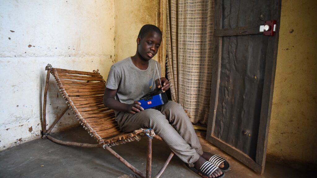 A young boy sits in a wooden chair at home, holding a radio.